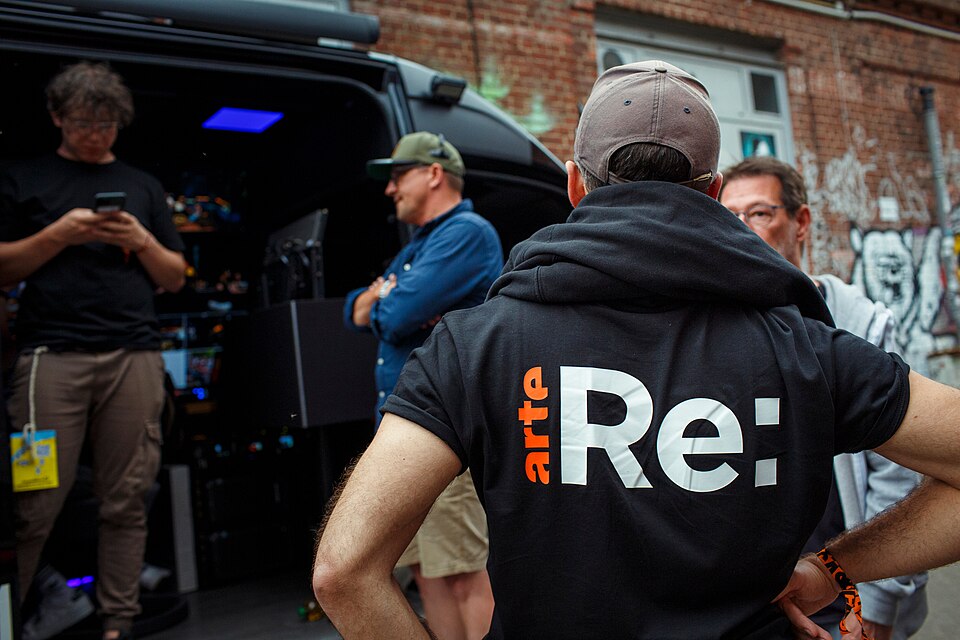 Man met pet op rug gezein met t-shirt Arte RE:, mannen op achtergrond
